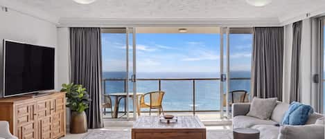 A modern living room with a large TV, a wooden coffee table, a sofa, and a view of the ocean through large sliding doors.