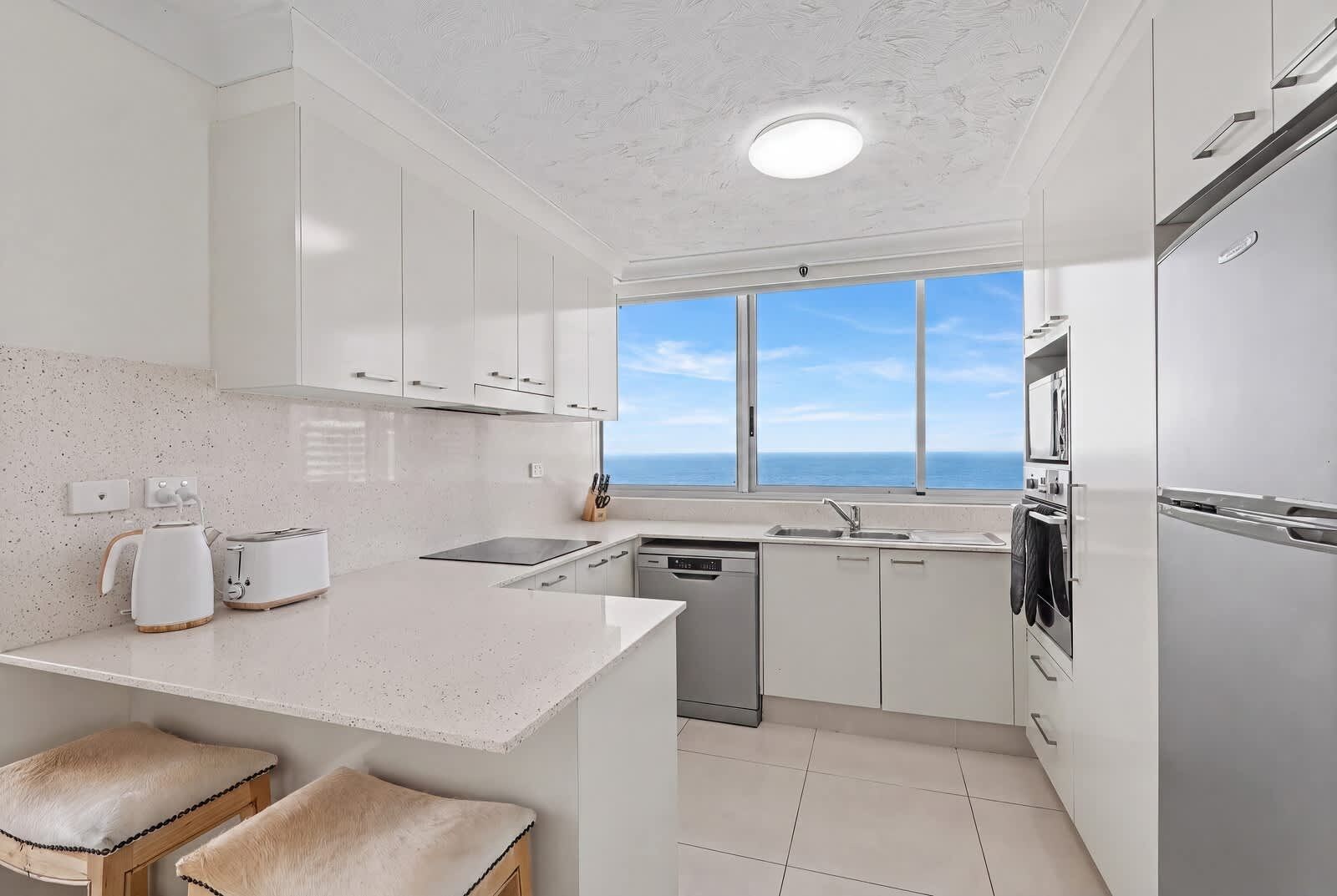 A modern kitchen with white cabinetry, a built-in oven, a dishwasher, and a view of the sea through large windows.