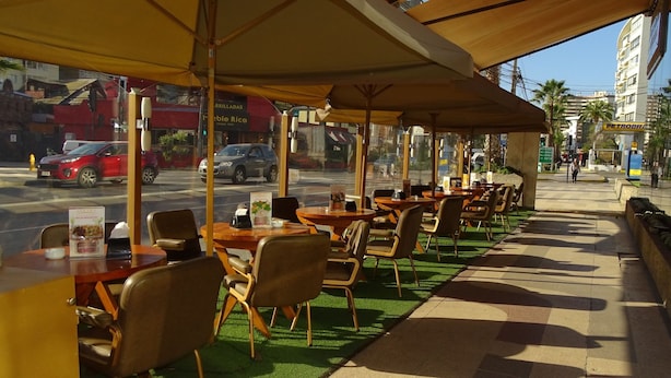 An outdoor cafe with tables and chairs under a canopy, overlooking a street with cars and buildings. - Hotel Ankara (Vina del Mar)