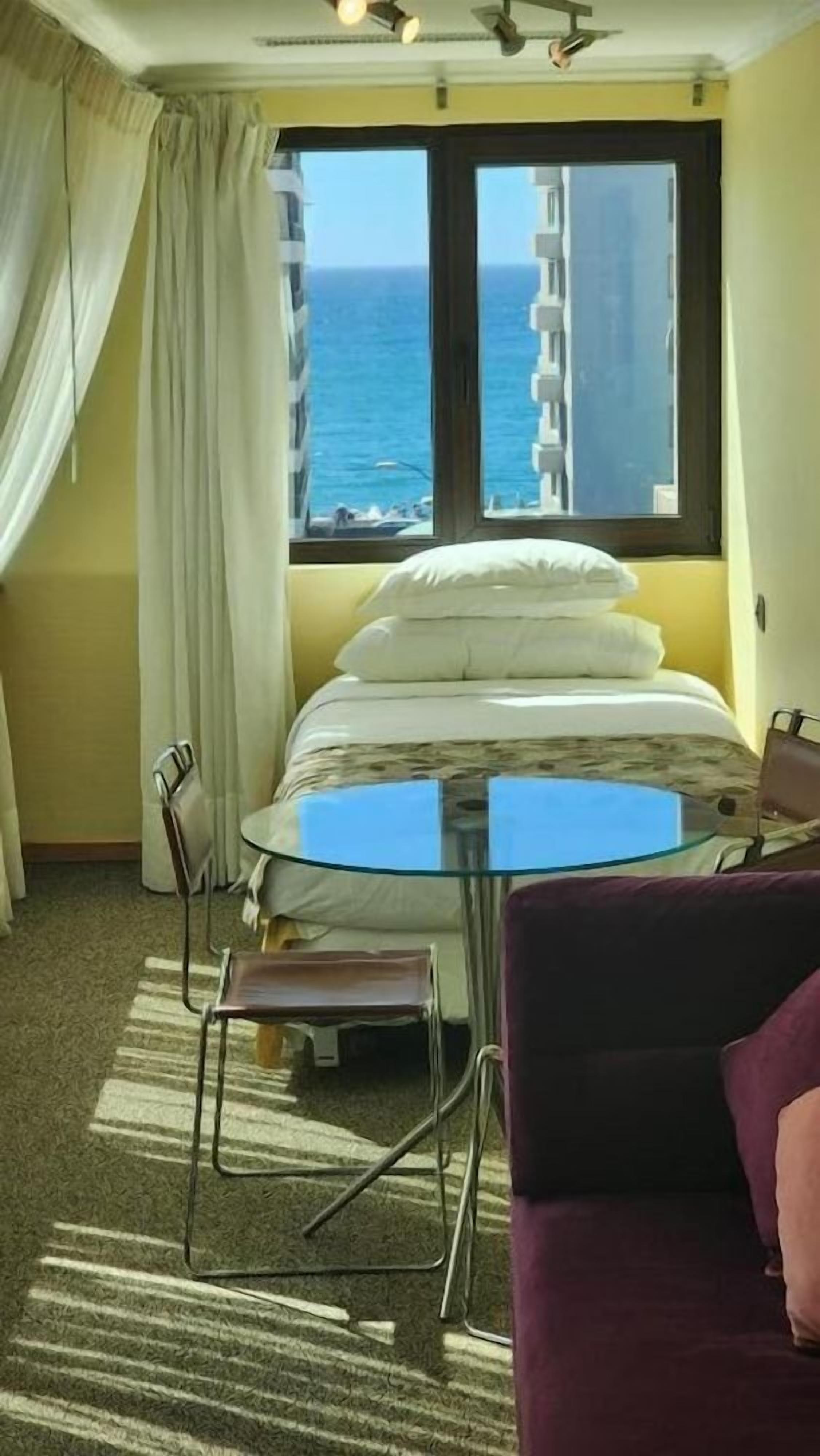 Une chambre d’hôtel avec un lit, une table en verre, une chaise et une vue sur l’océan.