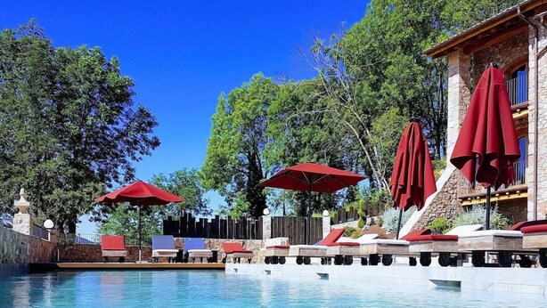 Ein klares Schwimmbecken mit Liegestühlen und roten Sonnenschirmen, umgeben von Bäumen und einem Steinhaus.