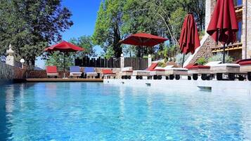 Una piscina azul cristalina con sillas de descanso y sombrillas rojas, rodeada de árboles y un edificio de piedra.