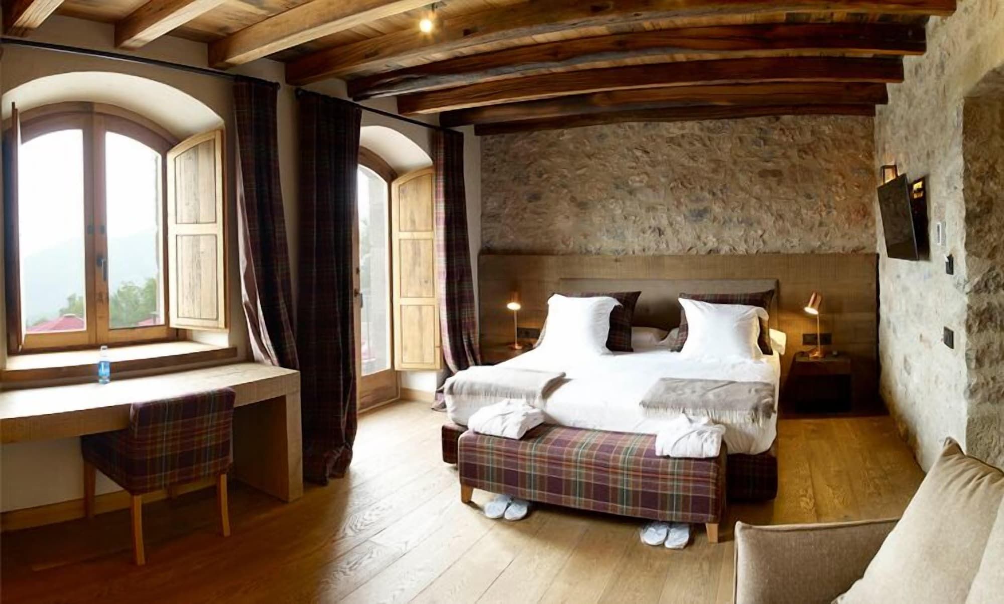 Un dormitorio con techo de madera, una cama grande con almohadas, una mesita de noche, una silla y una ventana con cortinas.