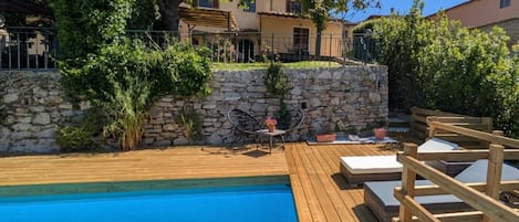 A swimming pool with a wooden deck, stone wall, and outdoor furniture.