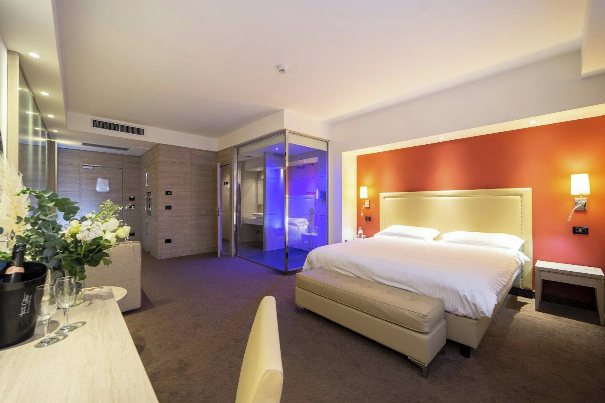 Une chambre d’hôtel comprenant un lit, un bureau avec une chaise, une petite table avec un vase de fleurs et une cabine de douche en verre.