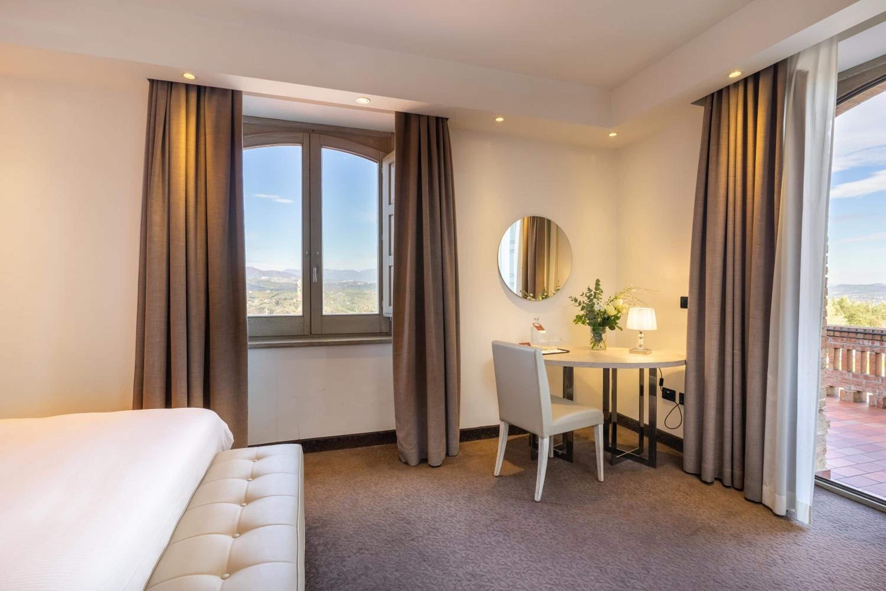 Habitación de hotel con cama, escritorio con silla, espejo redondo y vistas al exterior.
