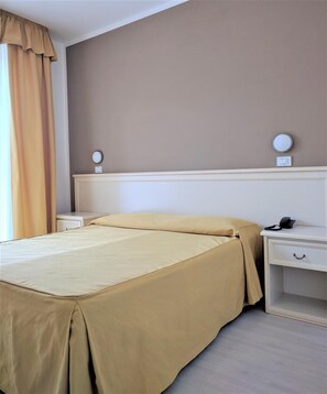 Una habitación de hotel con una cama, mesitas de noche, una lámpara y un ventanal con cortinas.
