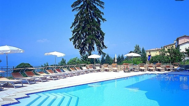 A swimming pool with sun loungers and umbrellas, surrounded by buildings and a tall tree. - Albergo Sole (San Zeno di Montagna)