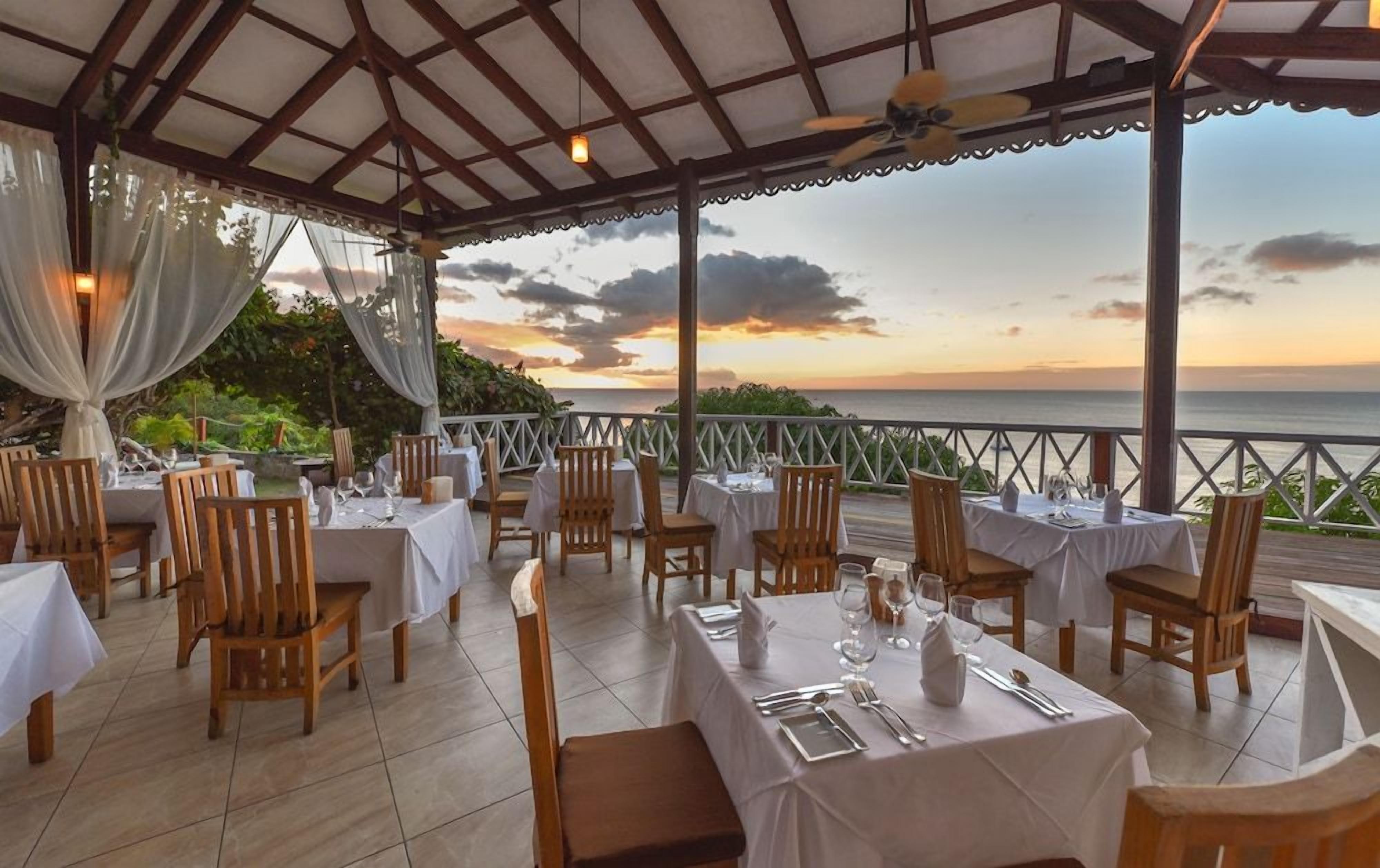 An outdoor dining area with tables and chairs, a view of the sea, and a covered structure with a ceiling fan.