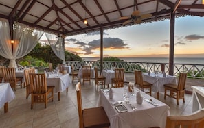 Un espace de restauration en plein air, avec des tables et des chaises, offrant une vue sur un plan d’eau au coucher du soleil.