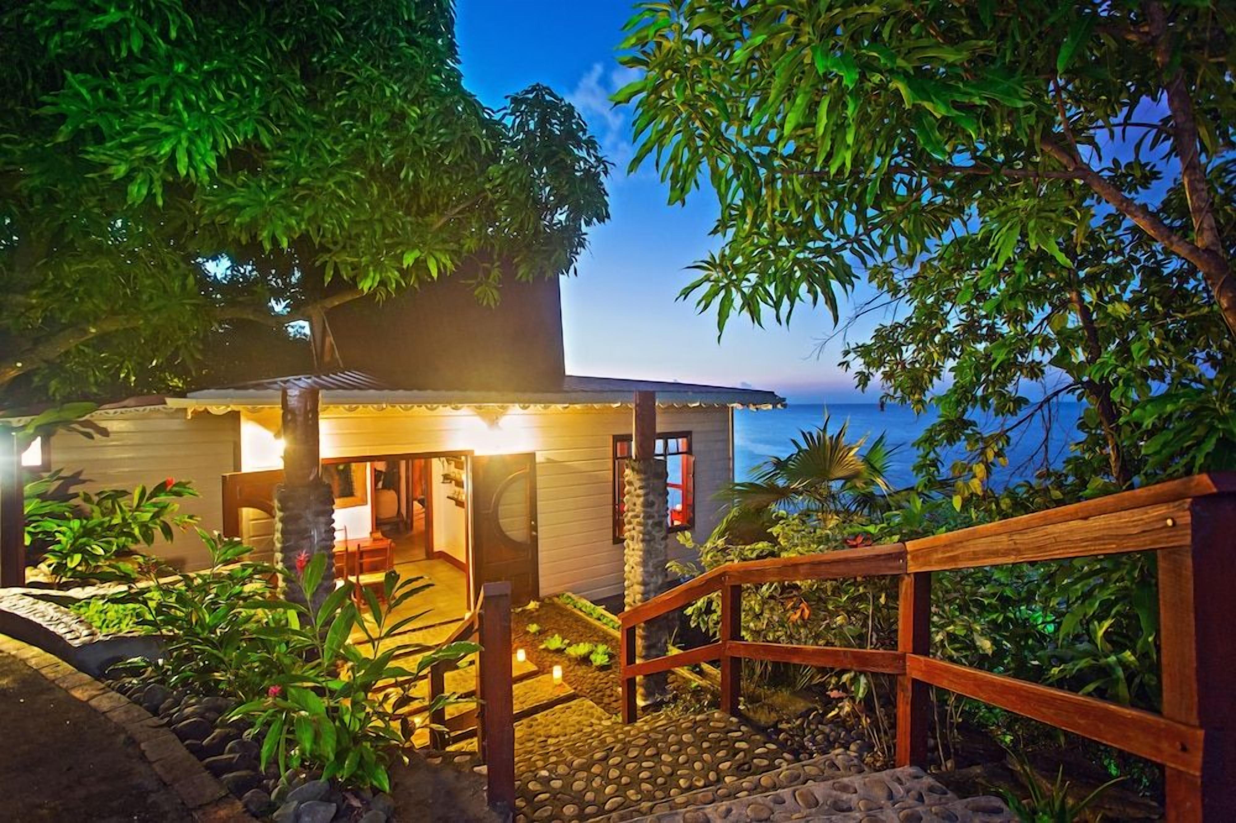 A wooden staircase leading to a house with a porch, surrounded by lush greenery and a view of the ocean.