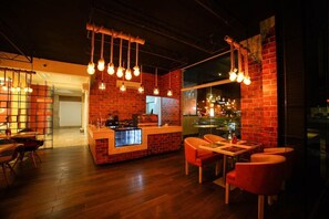 Interior de restaurante com paredes de tijolo, luminárias pendentes, área de bar e piso de madeira.