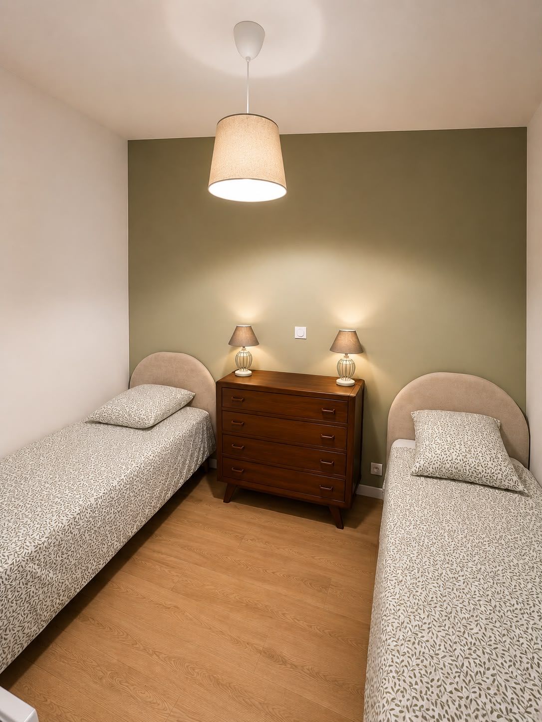 Quarto com duas camas de solteiro, cômoda de madeira e luminária pendente.