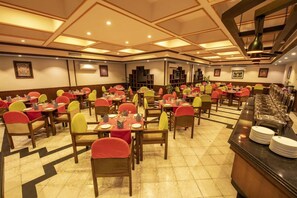 Uma sala de jantar com várias mesas e cadeiras, um balcão de buffet com pratos empilhados e um teto com iluminação embutida.