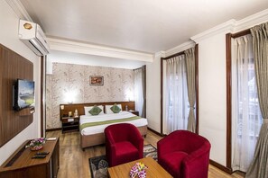 Quarto de hotel com cama, duas poltronas vermelhas, mesa de cabeceira de madeira, televisão e janela com cortinas.