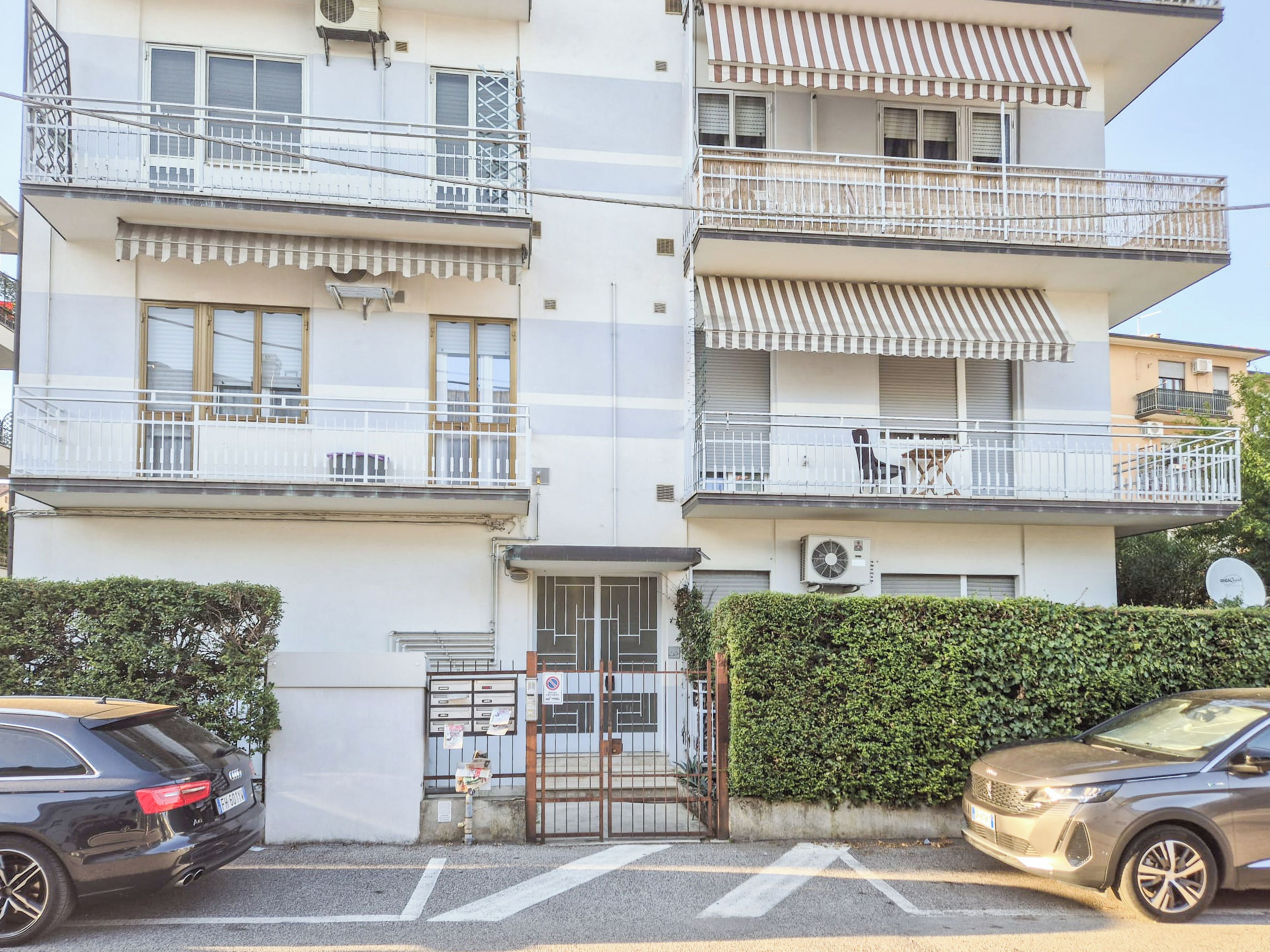 Edifício residencial com vários andares, varandas, portão fechado e carros estacionados.