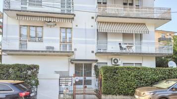 Edifício residencial com vários andares, varandas, portão fechado e carros estacionados.