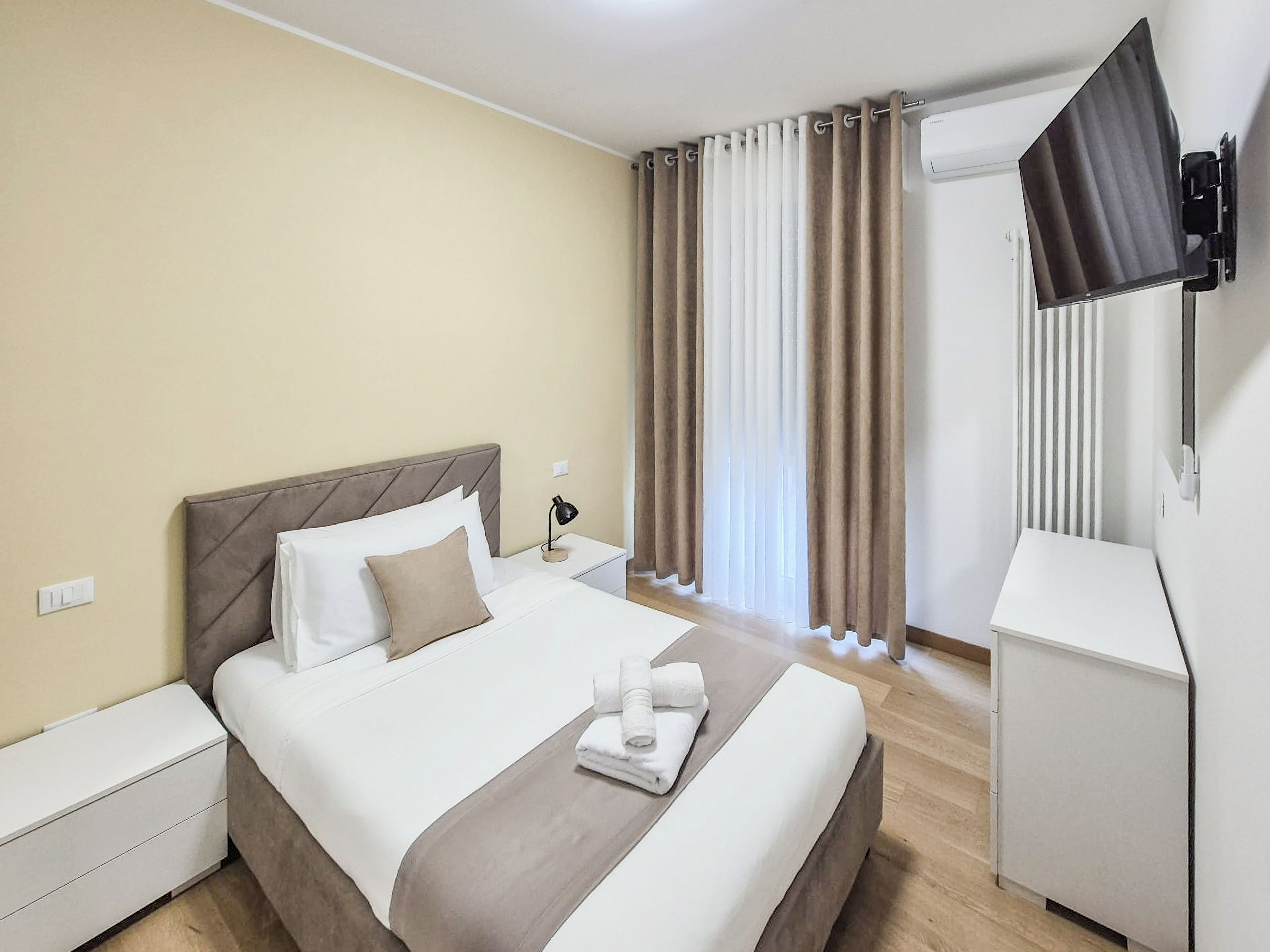 A hotel room with a bed, bedside tables, a flat-screen TV, and a window with curtains.