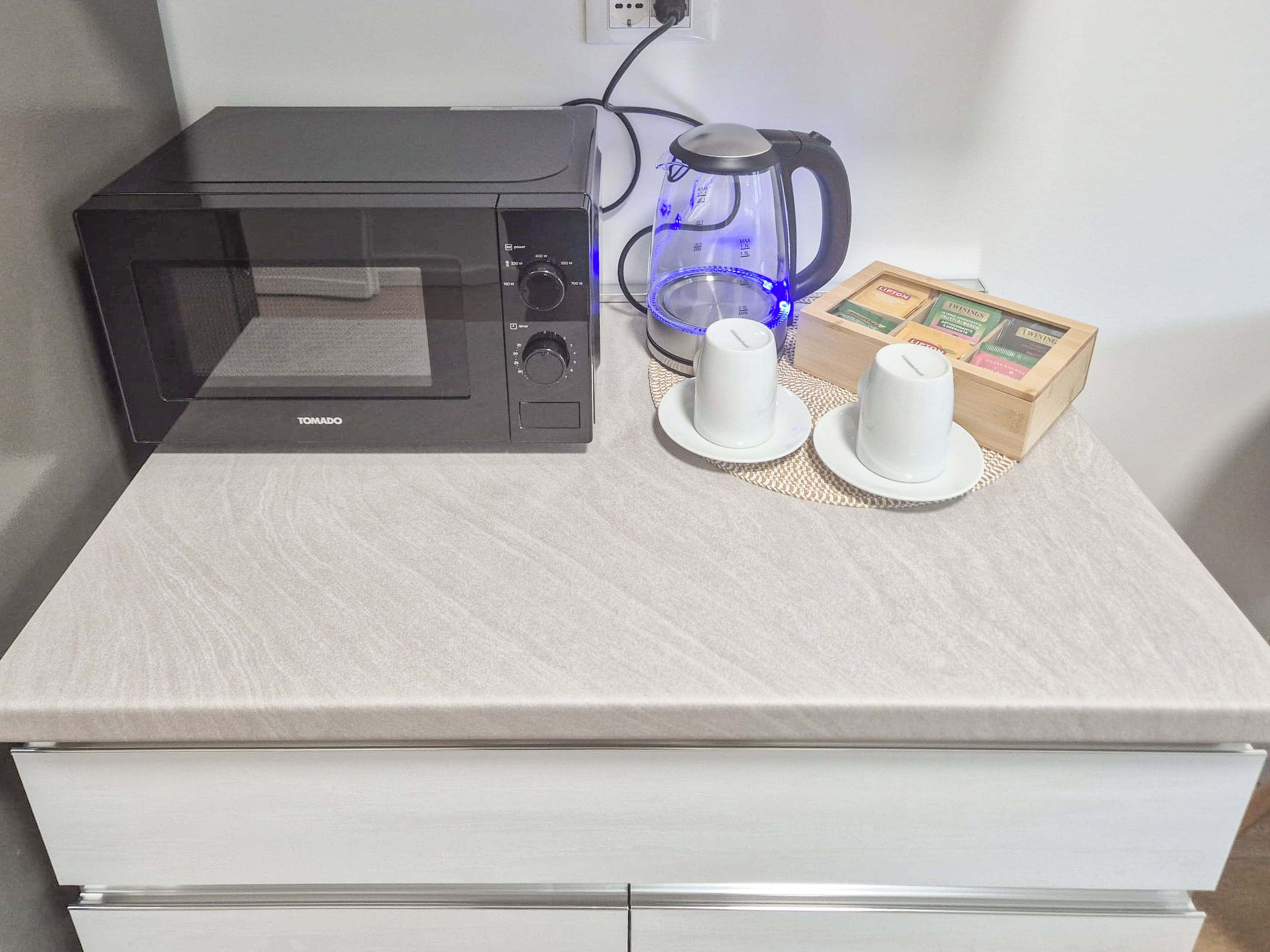 A kitchen area with a black microwave, a glass coffee maker with blue lights, and two white teacups on a woven placemat.