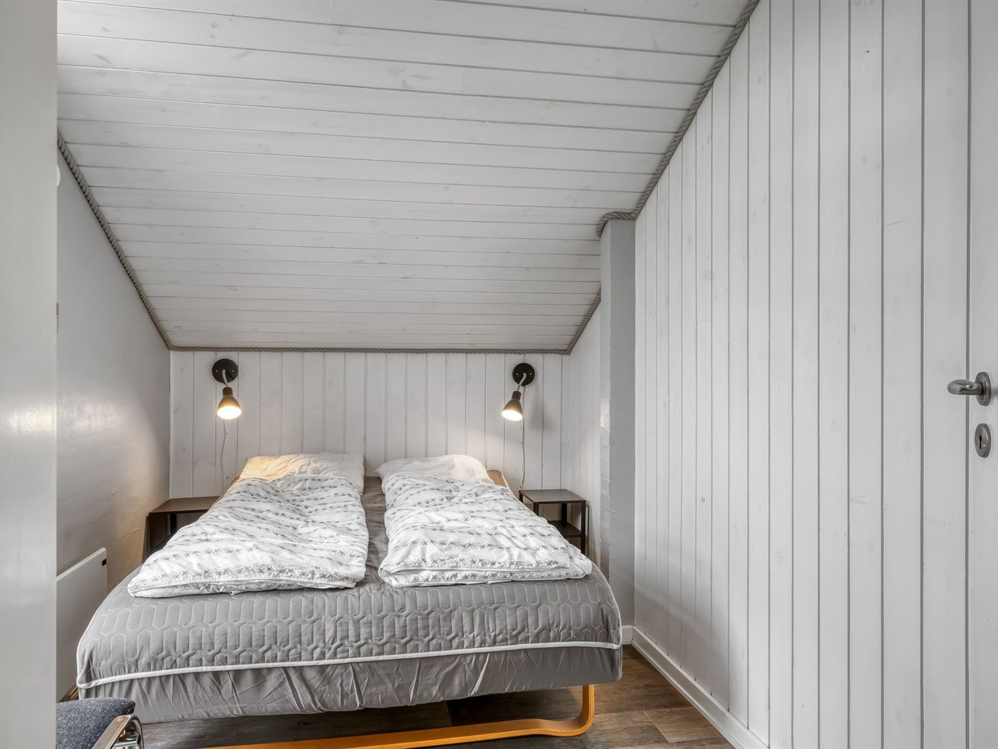A small, narrow room with a sloped ceiling and white wooden walls. There are two beds with grey bedding and two wall-mounted lamps.