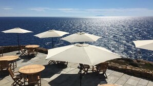 Ein Sitzbereich im Freien mit Tischen, Stühlen und Sonnenschirmen, mit Blick auf das Meer.