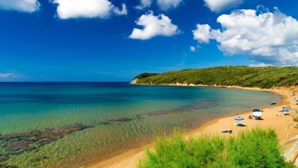 Ein Strand mit klarem türkisblauem Wasser, einem sandigen Ufer und einigen Sonnenschirmen.