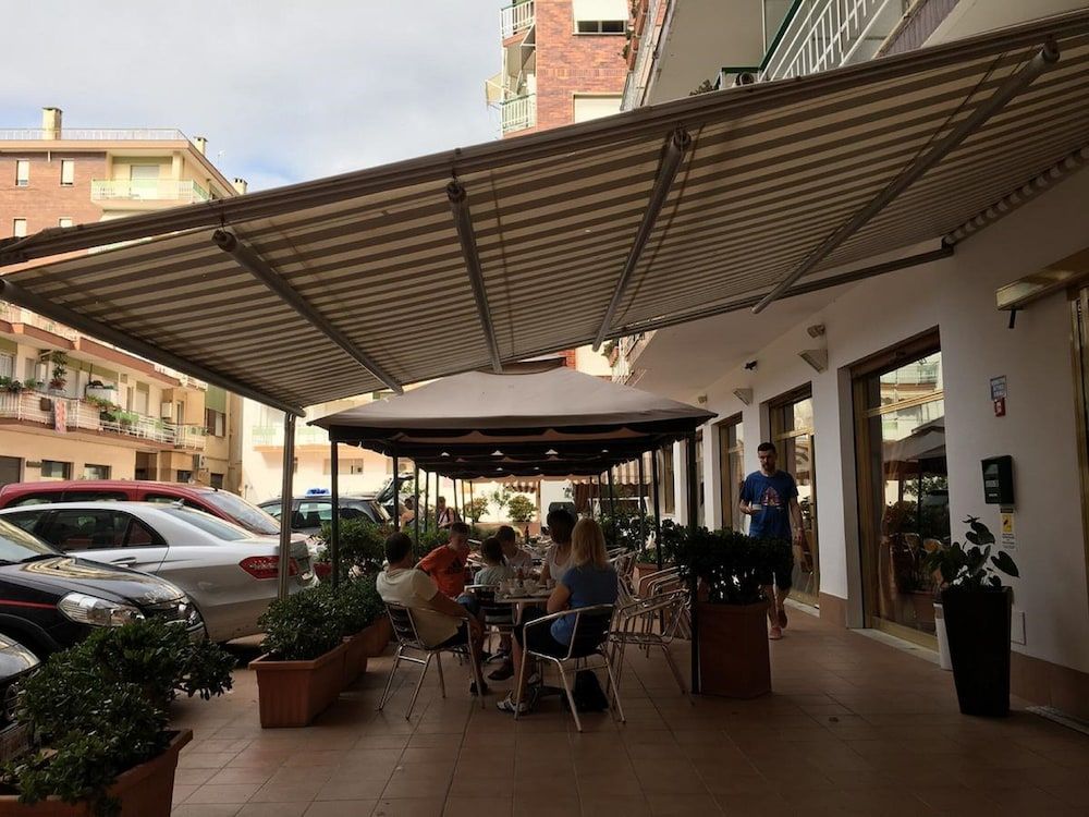 An outdoor cafe with tables and chairs under a covered area, surrounded by potted plants and parked cars.