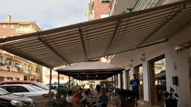 An outdoor cafe with tables and chairs under a striped awning, surrounded by potted plants and parked cars. - Arma (Taggia)