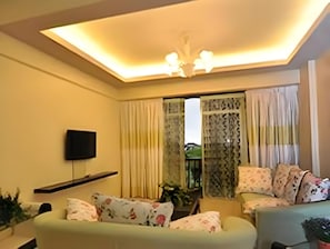 Uma sala de estar com sofá, TV fixada na parede e luminária no teto.