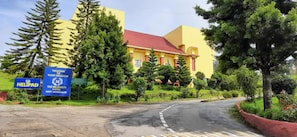 A yellow building with a red roof, surrounded by greenery and trees. There are signs indicating 'WELCOME TO YOUR KINGDOM' and 'THE MONARCH HOTEL.' - Howard Johnson Ooty India (Ootacamund)