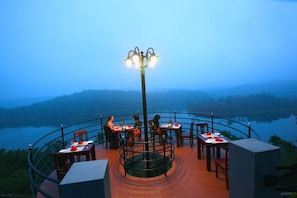 An outdoor dining area with tables and chairs arranged around a central light fixture, overlooking a body of water. - Arayal Resorts: A Unit of Sharoy Resort (Vythiri)