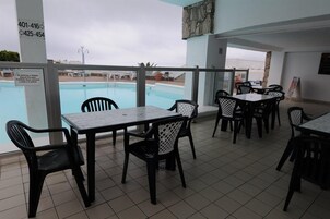 Área ao lado da piscina com mesas e cadeiras, vista da piscina e placa com números de contato de emergência.