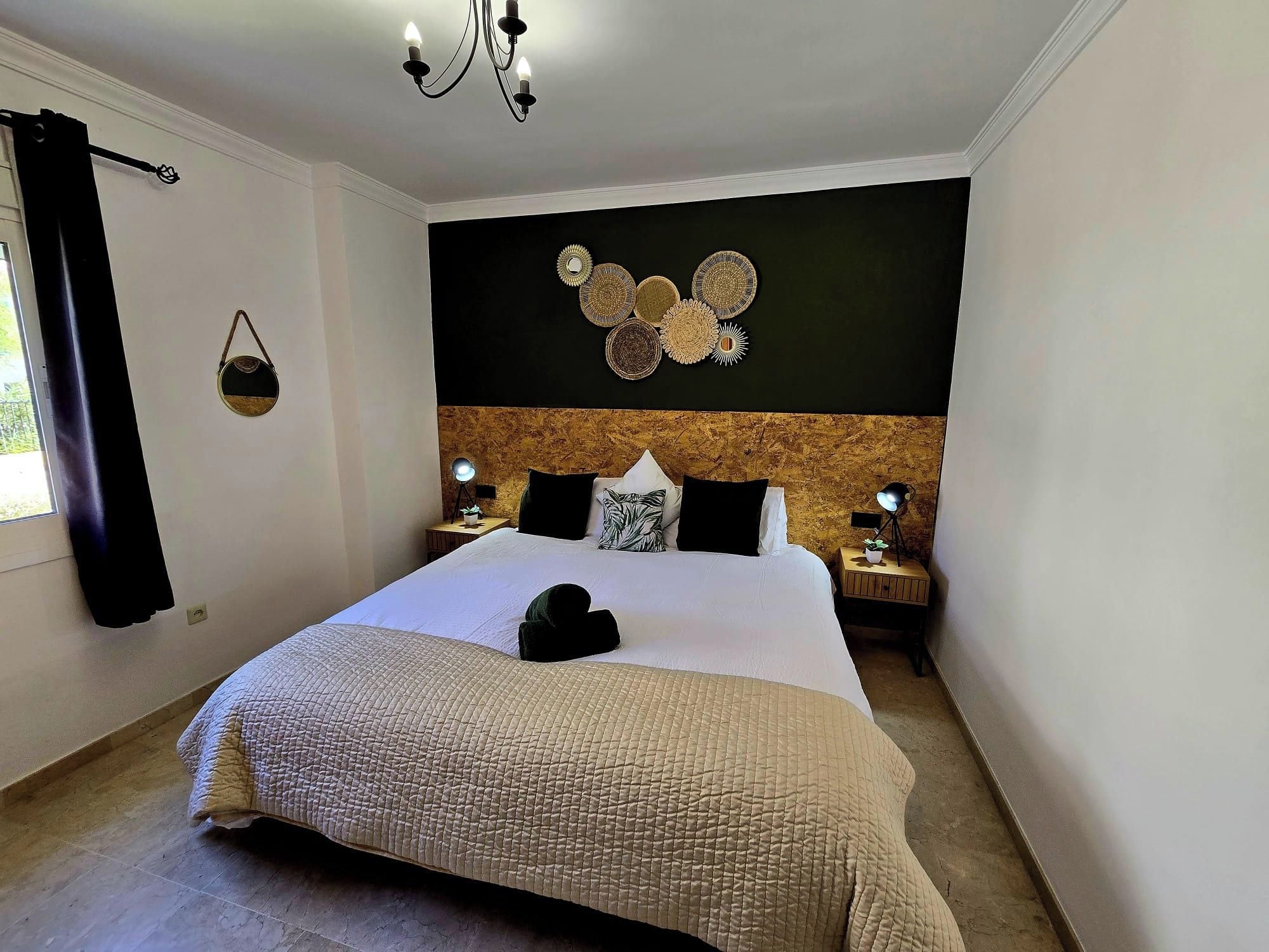 A hotel room with a large bed, two bedside tables with lamps, a hanging light fixture, a wall-mounted decorative piece, and a window with curtains.