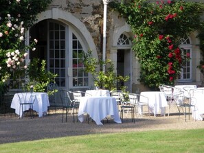 Un espace extérieur aménagé pour les repas, avec des tables et des chaises, un bâtiment en pierre doté de fenêtres en arc de cercle et des plantes en fleurs.