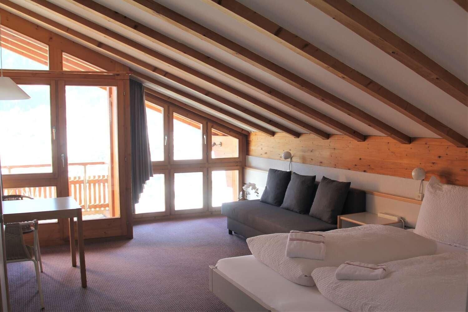 A room with a sloped ceiling, a bed, a sofa, a desk, and large windows.