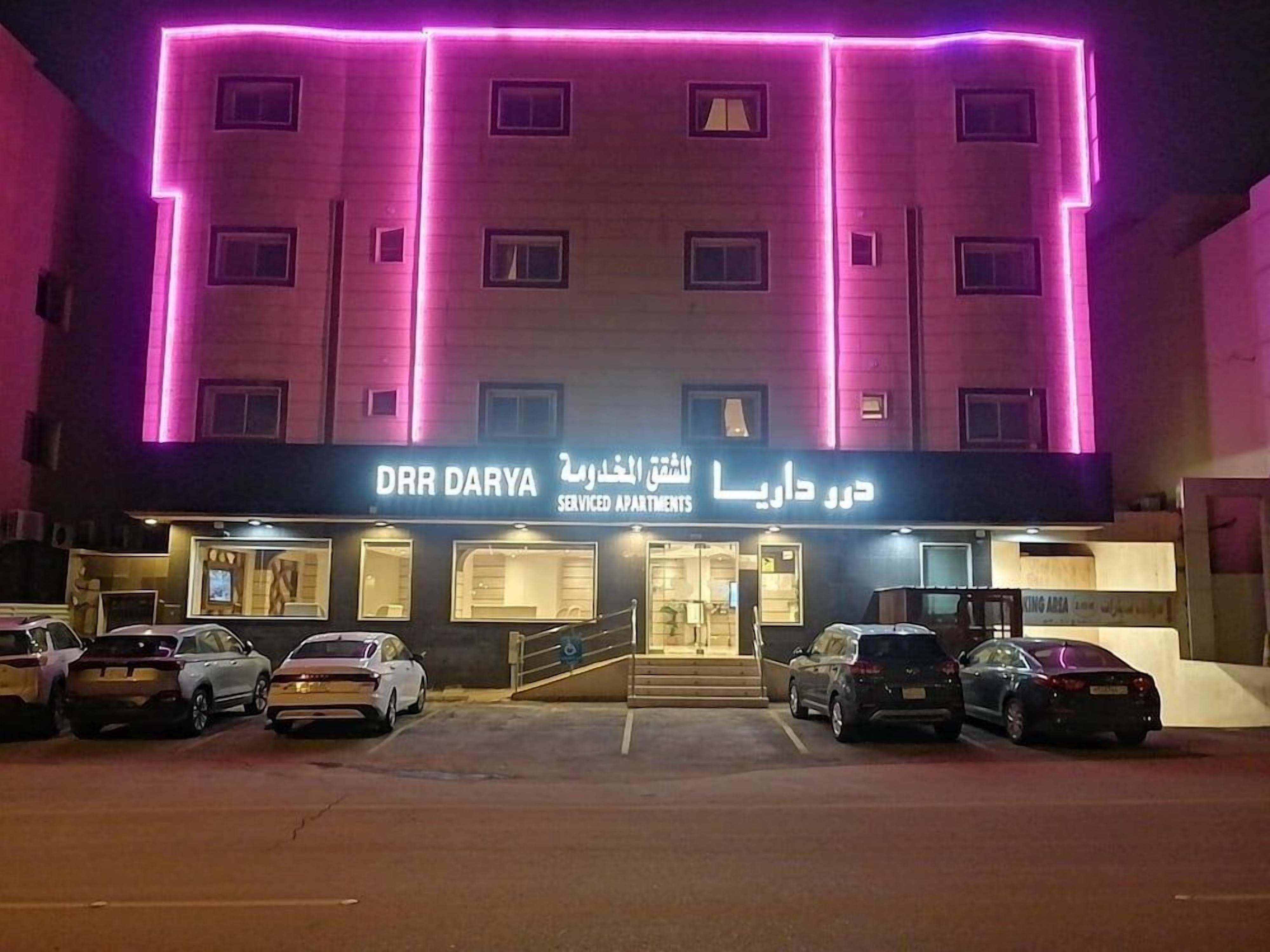 Un edificio con un letrero que dice 'DHR DARYA SERVICED APARTMENTS' y un estacionamiento con varios coches.