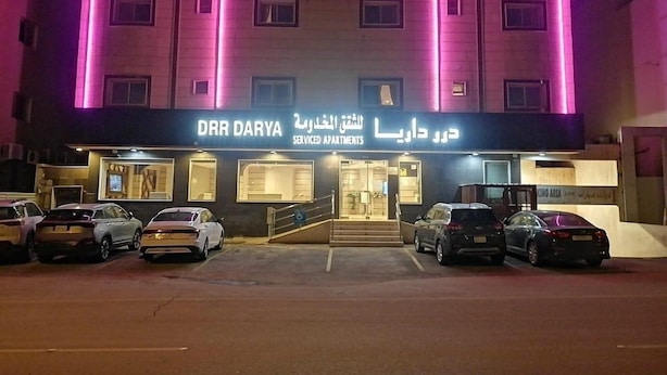 Um edifício com a placa 'DHR DARYA Serviced Apartments' e um estacionamento com vários carros.