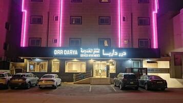 Un edificio con un letrero que dice 'DHR DARYA SERVICED APARTMENTS' y un estacionamiento con varios coches.