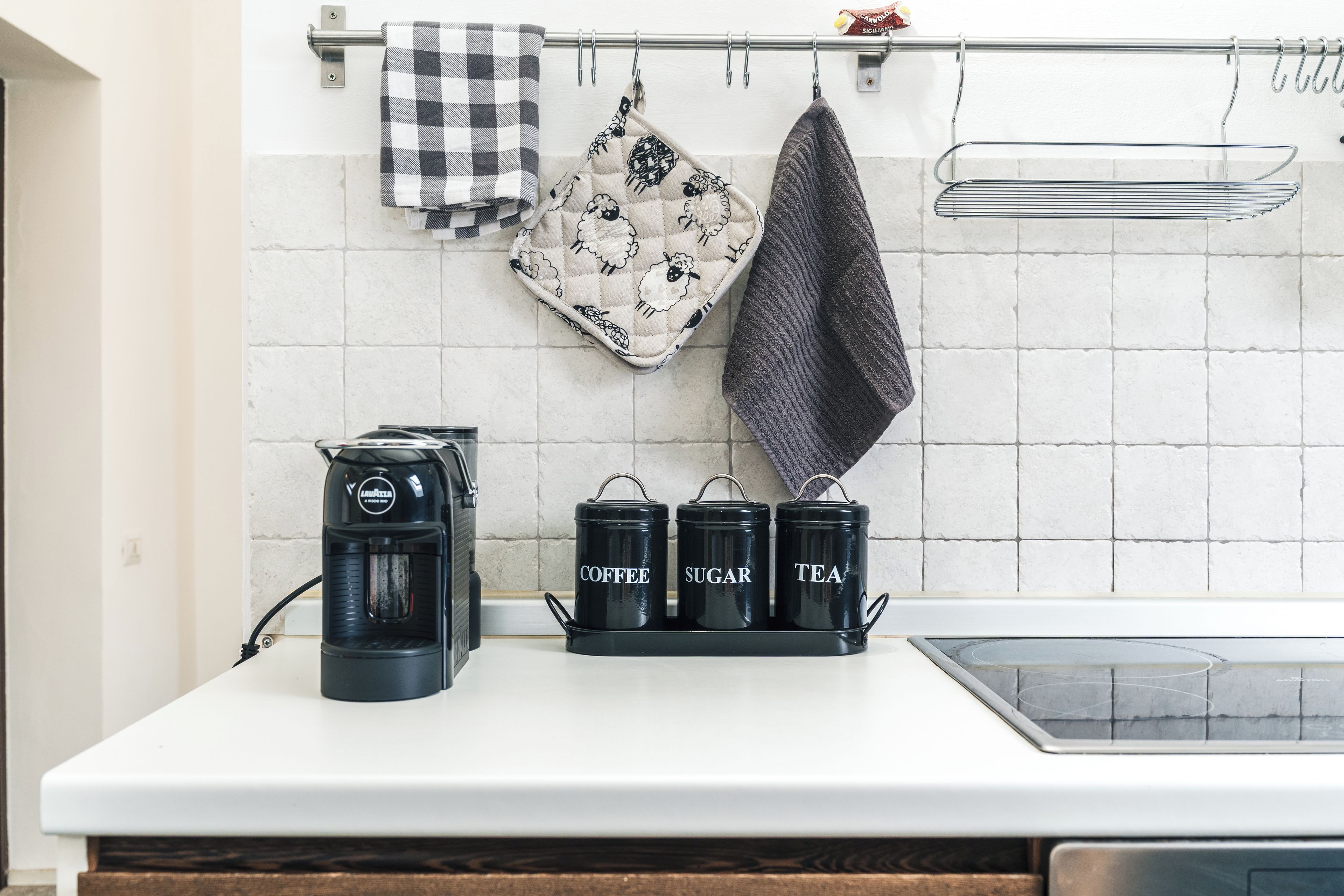 Eine Küchenarbeitsplatte mit einer Kaffeemaschine, zwei Behältern mit den Aufschriften 'COFFEE', 'SUGAR' und 'TEA' sowie einem Hängegestell mit Geschirrtüchern.