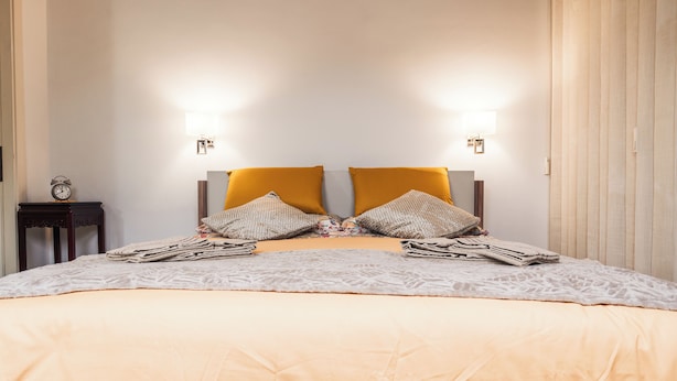 A neatly made bed with a patterned bedspread and two pillows, a bedside table with a clock, and wall-mounted lights. - Vrbo Property (Torino)