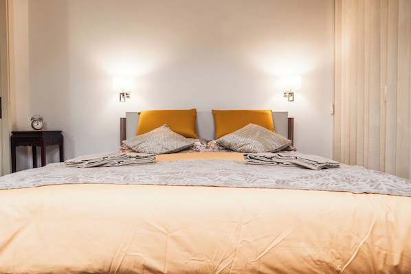 Un letto rifatto con cura, una coperta a fantasia e due cuscini, un comodino con una sveglia e luci montate a parete.