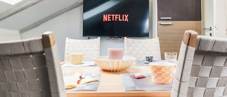 A dining area with a table set for two, a television displaying 'NETFLIX' on the screen, and a view of the sky through a skylight.