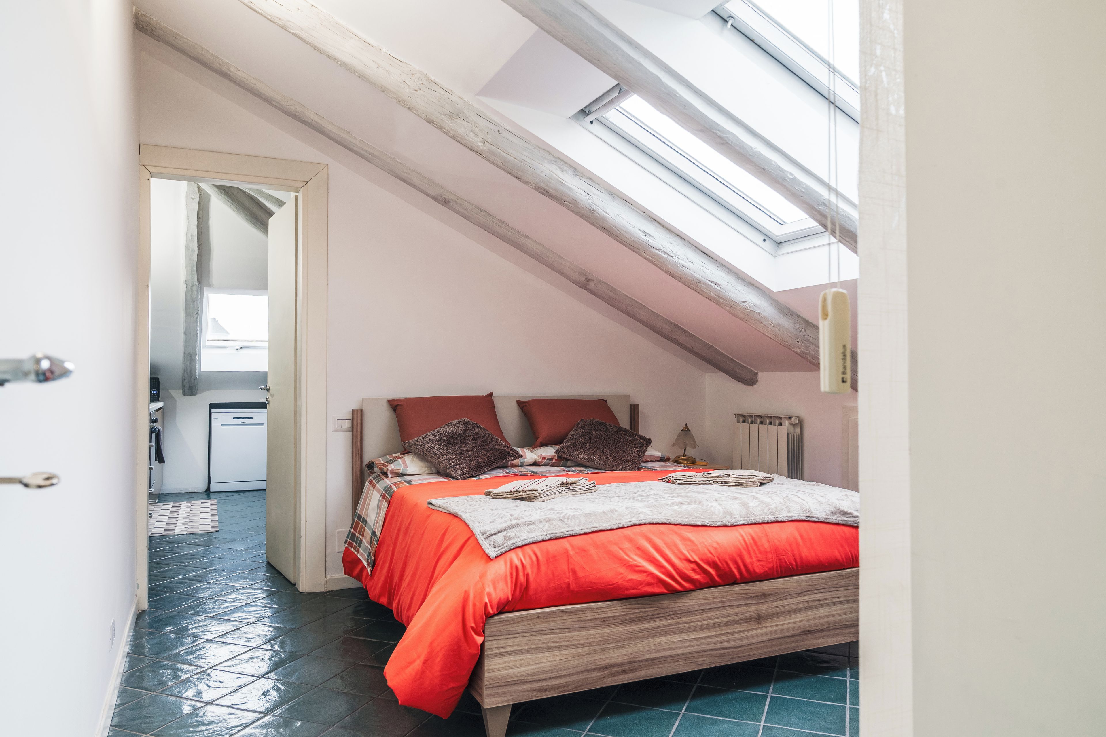 A bedroom with a sloped ceiling, a skylight, a bed with red bedding, and a visible kitchen area.
