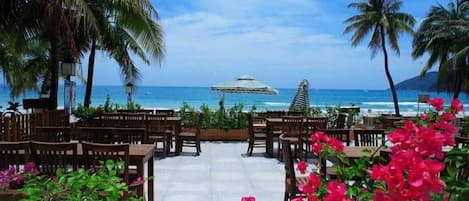 An outdoor seating area with wooden tables and chairs, palm trees, and a view of the ocean.