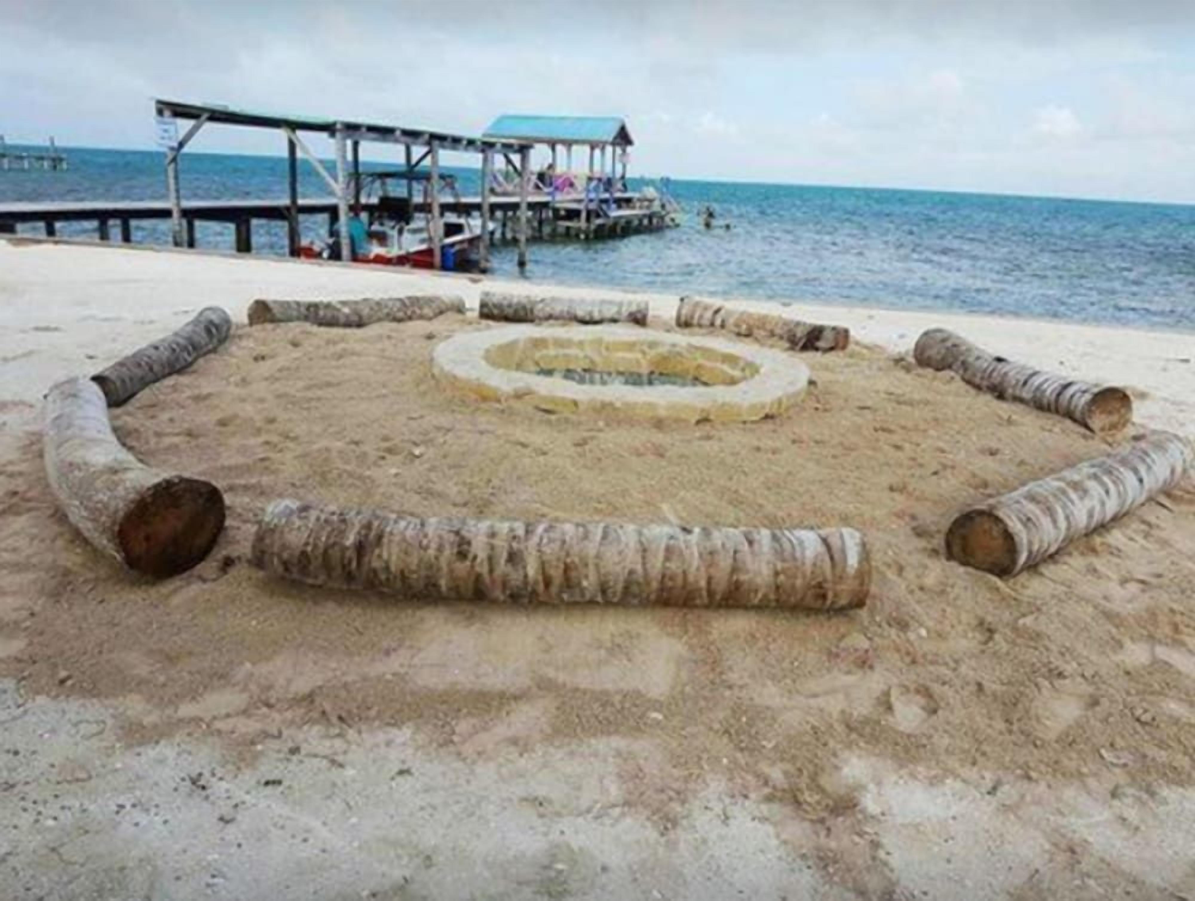 A sandy area with a circular stone structure, a wooden pier, and a body of water in the background.