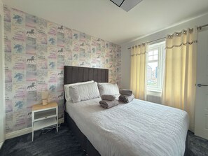 Ein Schlafzimmer mit einem Bett, Kissen, einem Nachttisch, einem Fenster mit Vorhängen und einer Wand mit einem sich wiederholenden Muster.