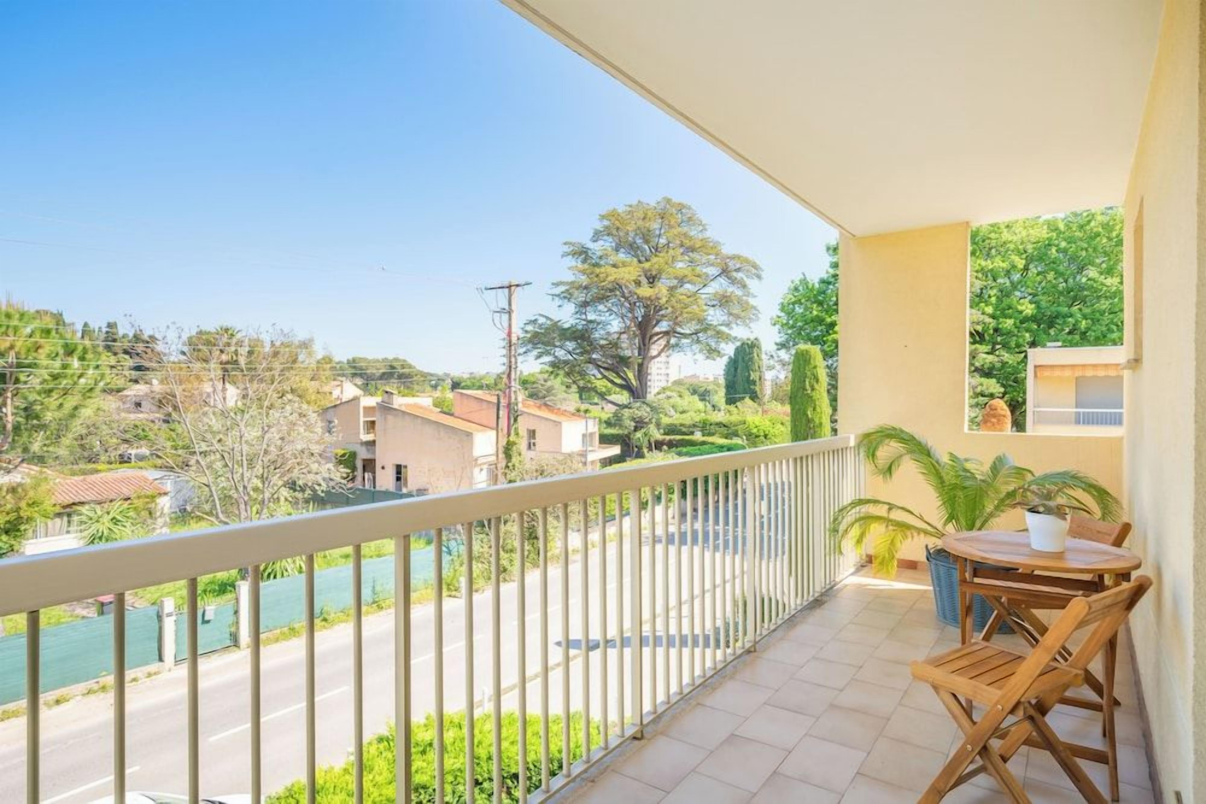 Ein Balkon mit Blick auf eine Straße und Grünflächen, ein Tisch mit Stühlen und ein Blumentopf.