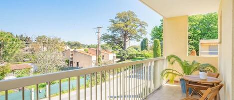 Ein Balkon mit Blick auf eine Straße und Grünflächen, ein Tisch mit Stühlen und ein Blumentopf.