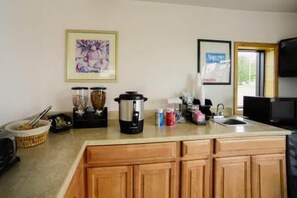 Cozinha com armários de madeira, cafeteira, chaleira e micro-ondas. Há um quadro na parede e uma janela com vista para o verde lá fora.