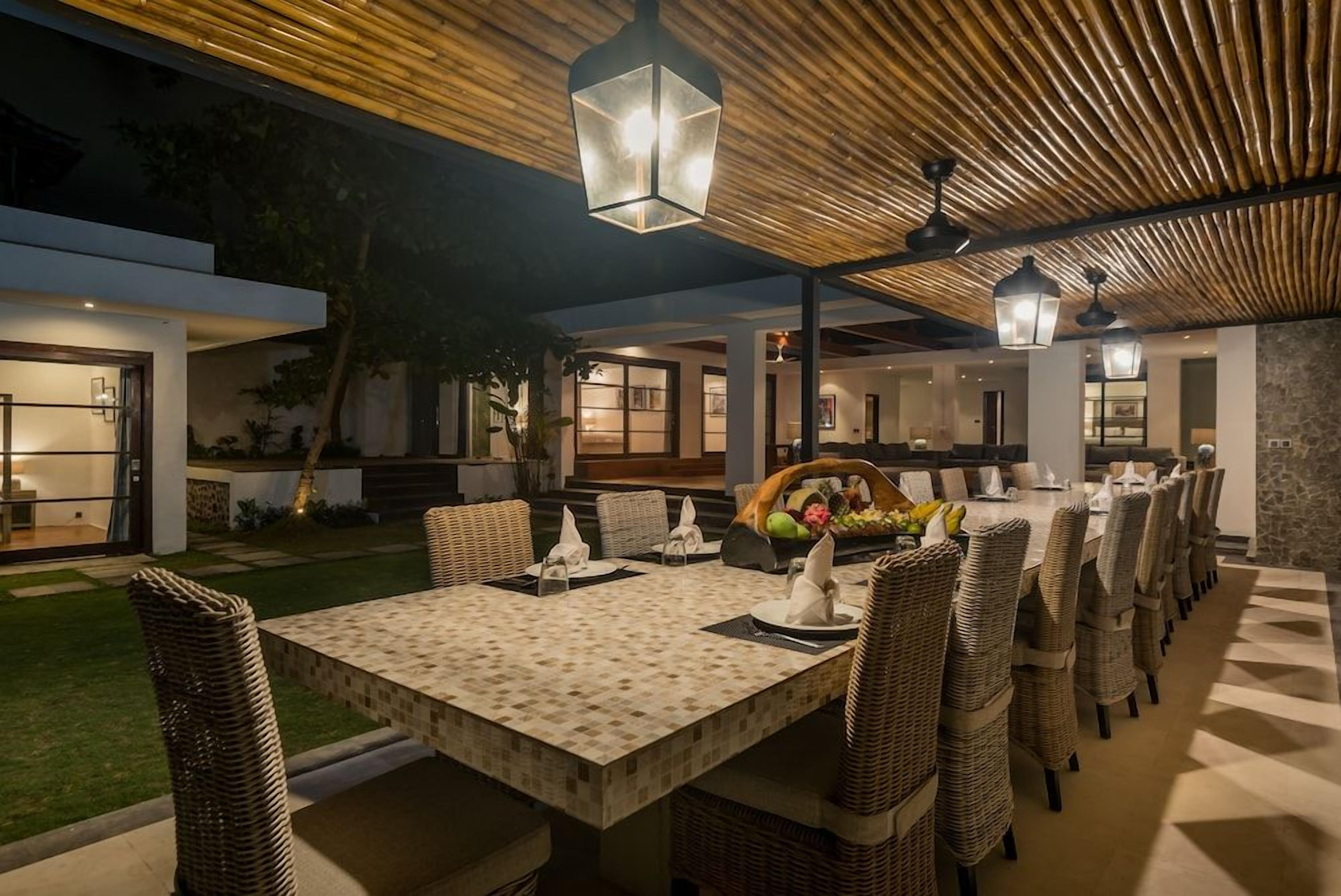 An outdoor dining area with a long table and wicker chairs, under a wooden ceiling with hanging lanterns.